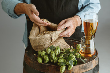 Close up of confident senior man brewer with self crafted beer in glass on wooden barrel on grey background. Owner of factory presented his products of brewing. Oktoberfest, drink, alcohol, industry.