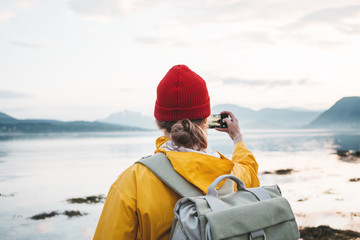 Wall Mural - Man traveler wearing yellow raincoat and backpack takes a photo on smartphone of great nordic landscape. Hipster tourist taking photo by smartphone trip summer vacations