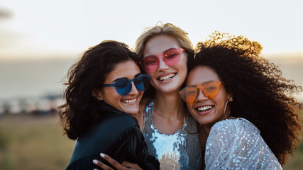 Wall Mural - Three happy women wearing sunglasses hugging at evening outdoors