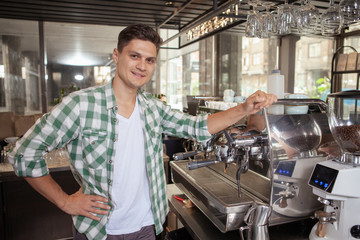 Charming friendly male barista smiling to the camera posing proudly at his coffee shop. Handsome coffee shop owner smiling to the camera, standing near coffee machine. Startup, business concept