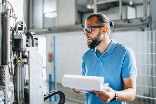 Fotomural Young engeneer manager with beard checking manufactory, workplace and machinery on big factory