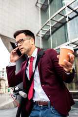 Wall Mural - Serious young businessman with electric scooter standing in front of modern business building talking at phone.