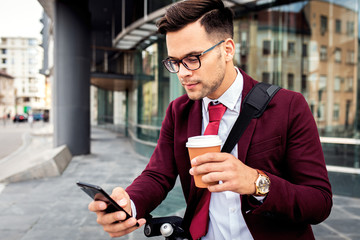 Wall Mural - Serious young businessman with electric scooter standing in front of modern business building looking at phone.