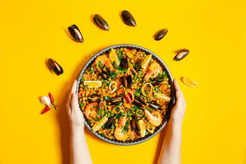 Hands holding pan with spanish paella with seafood on a yellow background