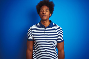 African american man with afro hair wearing striped polo standing over isolated blue background with serious expression on face. Simple and natural looking at the camera.