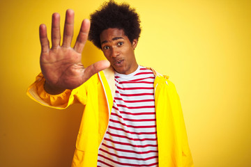 Canvas Print - African american man with afro hair wearing rain coat standing over isolated yellow background doing stop sing with palm of the hand. Warning expression with negative and serious gesture on the face.