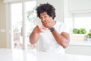 Sticker - Young african american man wearing casual white t-shirt sitting at home laughing at you, pointing finger to the camera with hand over mouth, shame expression