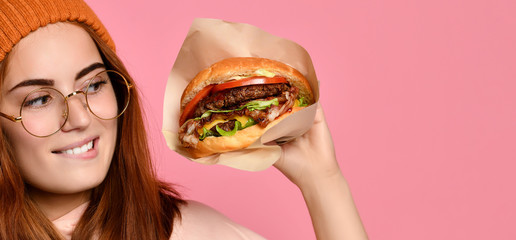 Beautiful teenage girl with red hair and hat holding burger and beverage in both hands. Ginger student girl has fast food lunch