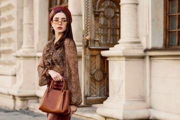 Wall Mural - Outdoor fashion portrait of young elegant, luxury lady wearing faux leather beret, skirt, leopard print blouse, beige glasses, holding brown bag, posing in street of European city. Copy, empty space