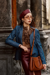 Outdoor autumn fashion portrait of young elegant, luxury woman wearing red faux leather beret, skirt, wrist watch, blue denim blazer, beige glasses, with brown bag, posing in street of European city