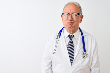 Canvas Print - Senior grey-haired doctor man wearing stethoscope standing over isolated white background puffing cheeks with funny face. Mouth inflated with air, crazy expression.