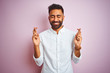 Young indian businessman wearing elegant shirt standing over isolated pink background gesturing finger crossed smiling with hope and eyes closed. Luck and superstitious concept.