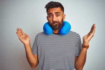 Wall Mural - Young indian tourist man on travel wearing neckpillow over isolated white background clueless and confused expression with arms and hands raised. Doubt concept.