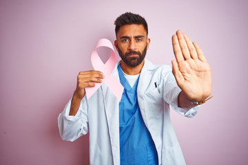 Poster - Young indian doctor man holding cancer ribbon standing over isolated pink background with open hand doing stop sign with serious and confident expression, defense gesture