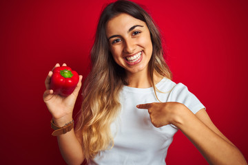 Sticker - Young beautiful woman holding pepper over red isolated background with surprise face pointing finger to himself