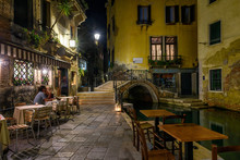 Narrow Canal With Bridge And Tables Of Restaurant In Venice, Italy. Architecture And Landmark Of Venice. Night Cozy Cityscape Of Venice.