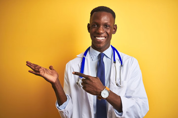 African american doctor man wearing stethoscope standing over isolated yellow background amazed and smiling to the camera while presenting with hand and pointing with finger.