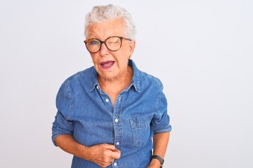 Senior grey-haired woman wearing denim shirt and glasses over isolated white background winking looking at the camera with sexy expression, cheerful and happy face.