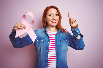 Sticker - Young beautiful redhead woman holding cancer ribbon over pink isolated background surprised with an idea or question pointing finger with happy face, number one