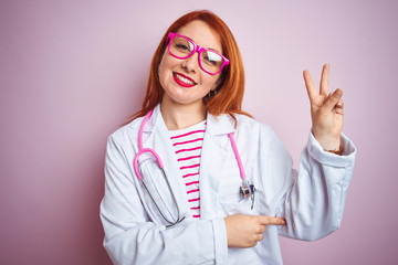 Poster - Young redhead doctor woman using stethoscope standing over isolated pink background smiling with happy face winking at the camera doing victory sign. Number two.