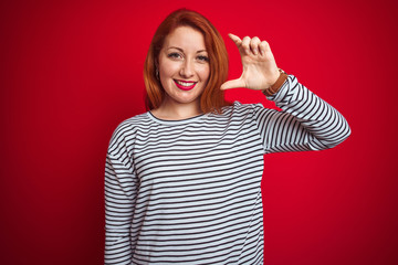 Wall Mural - Young redhead woman wearing strapes navy shirt standing over red isolated background smiling and confident gesturing with hand doing small size sign with fingers looking and the camera