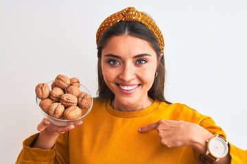 Wall Mural - Young beautiful woman holding bowl with walnuts standing over isolated white background with surprise face pointing finger to himself