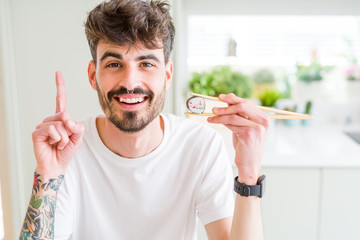 Sticker - Young man eating asian sushi using chopsticks surprised with an idea or question pointing finger with happy face, number one