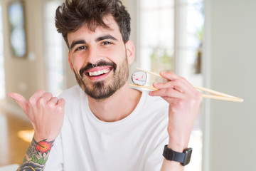 Sticker - Young man eating asian sushi using chopsticks pointing and showing with thumb up to the side with happy face smiling