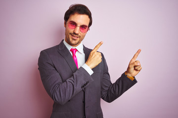 Sticker - Young handsome businessman wearing suit and sunglasses over isolated pink background smiling and looking at the camera pointing with two hands and fingers to the side.