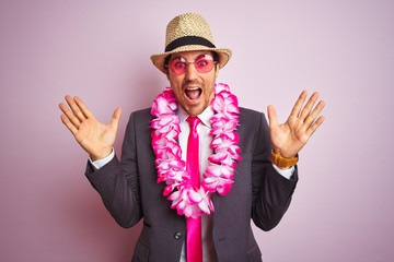 Young businessman wearing suit hat glasses hawaiian lei over isolated pink background very happy and excited, winner expression celebrating victory screaming with big smile and raised hands