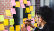 © weedezign - asian businessman looking and writing on sticky note for brainstorming ideas on clear whiteboard at modern office. analyse and synthesis
