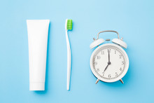 Alarm clock, toothbrush with green bristles and white tube of toothpaste on pastel blue background. Healthy teeth. Morning routine concept. 