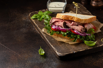 Wall Mural - Roast beef sandwich with tomato, onion, lettuce and mustard sauce on dark background. Delicious healthy lunch with meat, hearty food