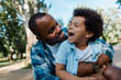 © LIGHTFIELD STUDIOS - happy african american man hugging positive kid in park