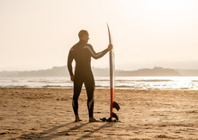 Rear View Of Strong Surfer With Surfboard On The Beach At Sunset Or Sunrise.