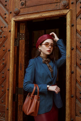 Wall Mural - Outdoor fashion portrait of young elegant, luxury woman wearing red faux leather beret, skirt, wrist watch, blue denim blazer, beige glasses, holding brown handbag, posing in street of European city