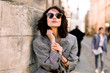 © sofiko14 - Beautiful brunette young woman wearing jacket and black sunglasses walking on the street, posing near the old building in ancient city