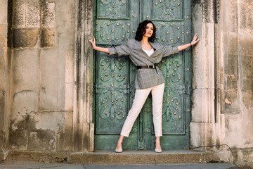 A young woman in white pants and checkered jacket with dark hair stands and looks into the distance on the background of an old building with vintage doors.