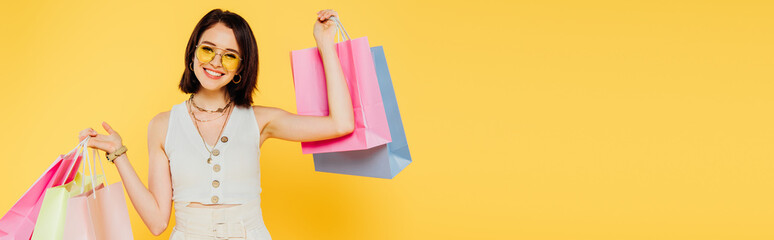 Wall Mural - panoramic shot of happy fashionable girl in sunglasses holding shopping bags isolated on yellow