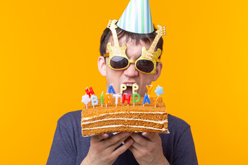 Wall Mural - Positive funny young asian guy with a cap and a burning candle and a homemade cake in his hands posing on a yellow background. Anniversary and birthday concept.