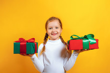 Portrait Of A Happy Little Girl In A White Turtleneck On A Yellow Background. The Child Holds Boxes With Gifts In His Hands
