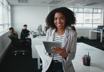 Wall Mural - Portrait of a smiling confident african american young businesswoman holding digital tablet in hand looking at camera with colleague at background in office