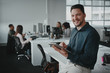 © StratfordProductions - Happy young professional businessman with digital tablet looking at camera against his coworker working in background