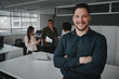 © StratfordProductions - Portrait of a happy successful young businessman with his arms crosses looking at camera and his colleague working in the office at background