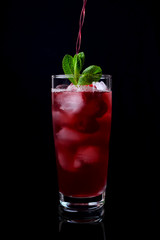 Process of pouring the red drink into a tall glass filled with ice cubes, raspberries and mint against the black background