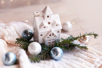 Wall Mural - Ceramic house, Christmas cozy decorations on wooden and knitted background with glares. Night lamp is decorated with blue and white ornaments, fir branch, new year lights, garland.