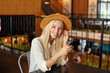 Beautiful young blonde woman with long hair wearing hat and white shirt, sitting at table over cafe interior, looking at camera with sincere smile