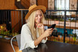 Positive attractive blonde woman with long hair keeping chin on her hand while looking at her smartphone, sitting over cafe interior in brown hat