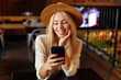 Indoor close-up of charming blonde young female in white shirt and brown hat sitting over cafe interior, holding mobile phone in hand and looking at screen with cheerful smile