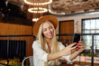 Beautiful young long haired blonde female in hat and white shirt, posing over cafe interior, smiling widely to camera while making selfie with her smartphone